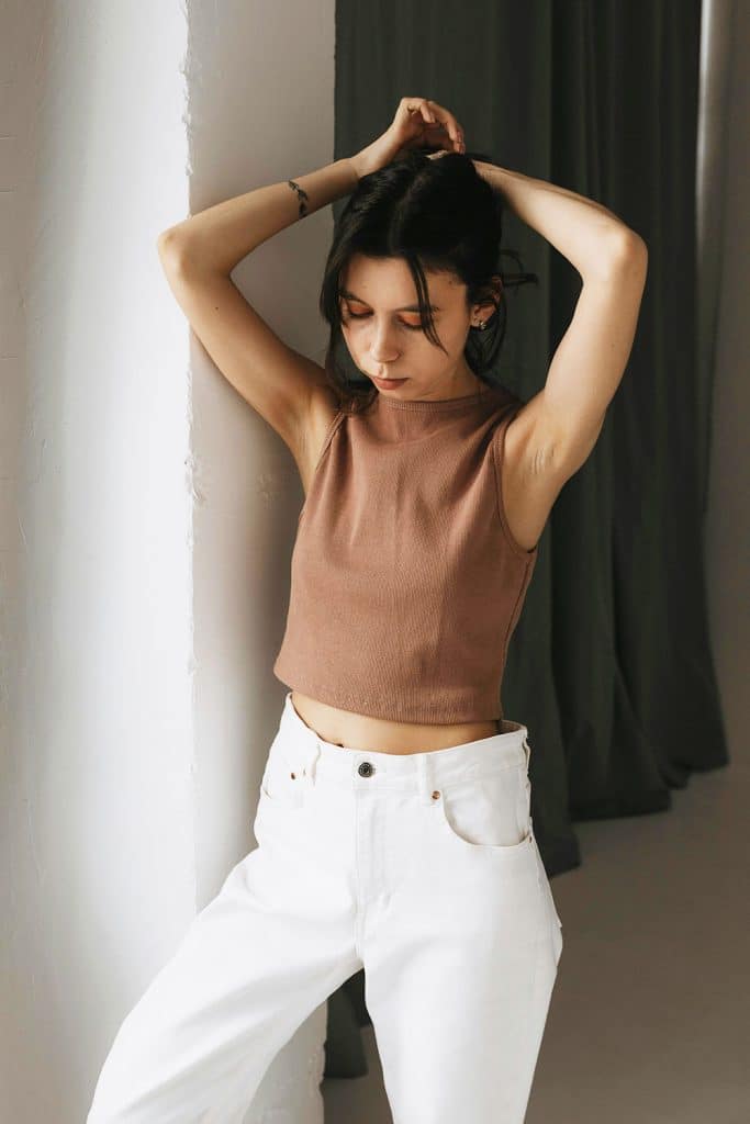 Woman in a brown sleeveless top and white pants stands with arms raised, eyes closed.