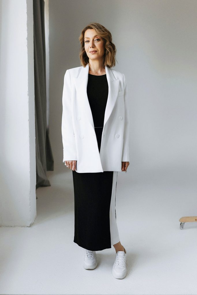 Woman in a white blazer, black dress, and white sneakers stands in a bright studio.