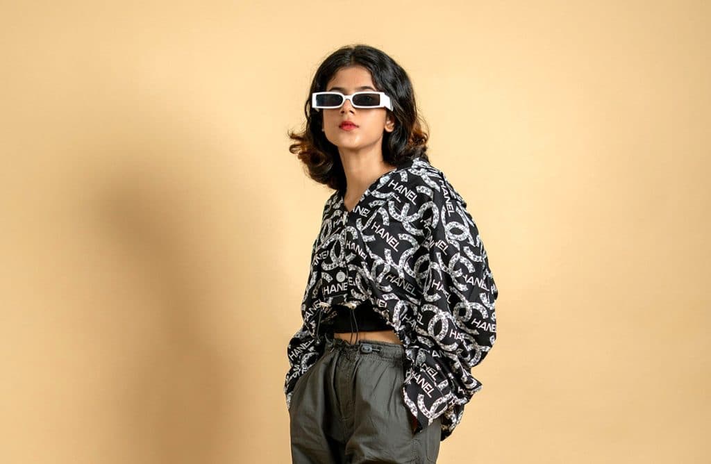 Woman in graphic shirt and gray pants poses against beige background, wearing white sunglasses.