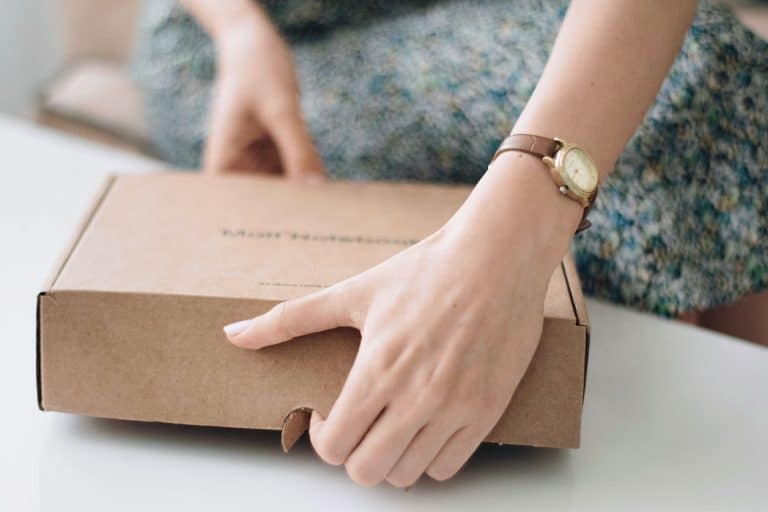A person wearing a watch opens a brown cardboard box on a white table.