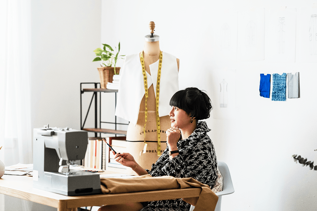 Fashion designer sits at a desk with a sewing machine, mannequin, and fabric samples.