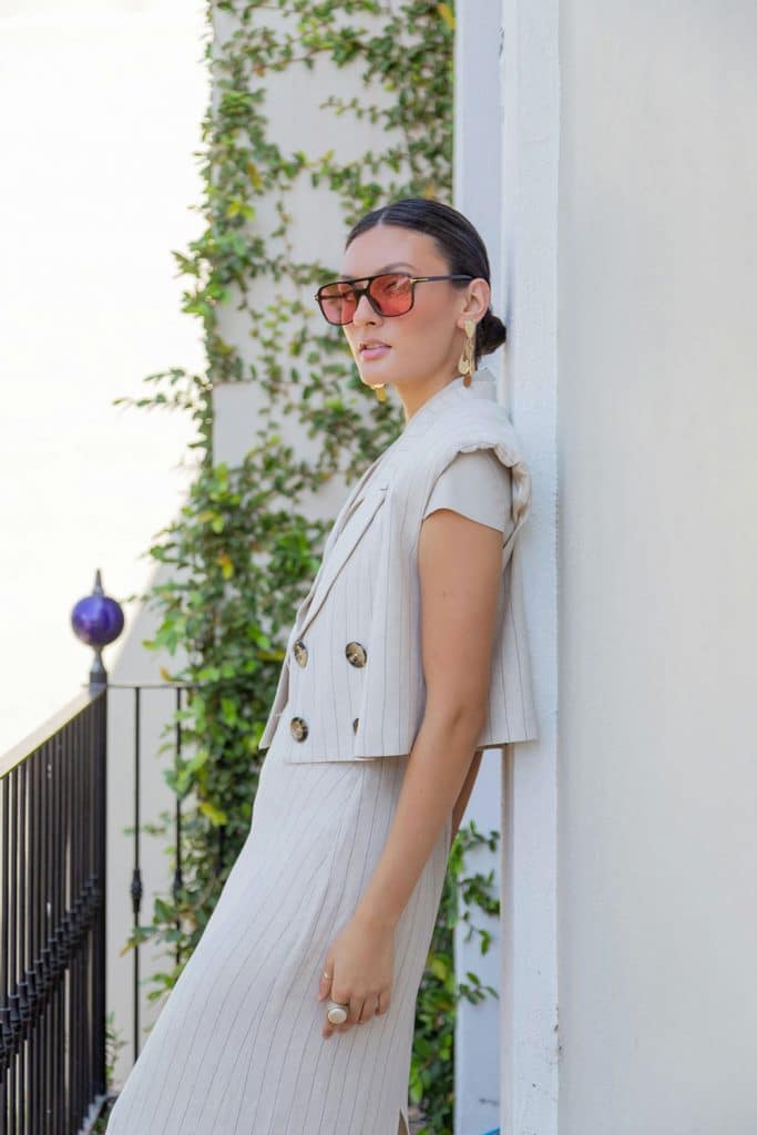 Woman in cream pinstripe suit and sunglasses leans against a white wall outdoors.