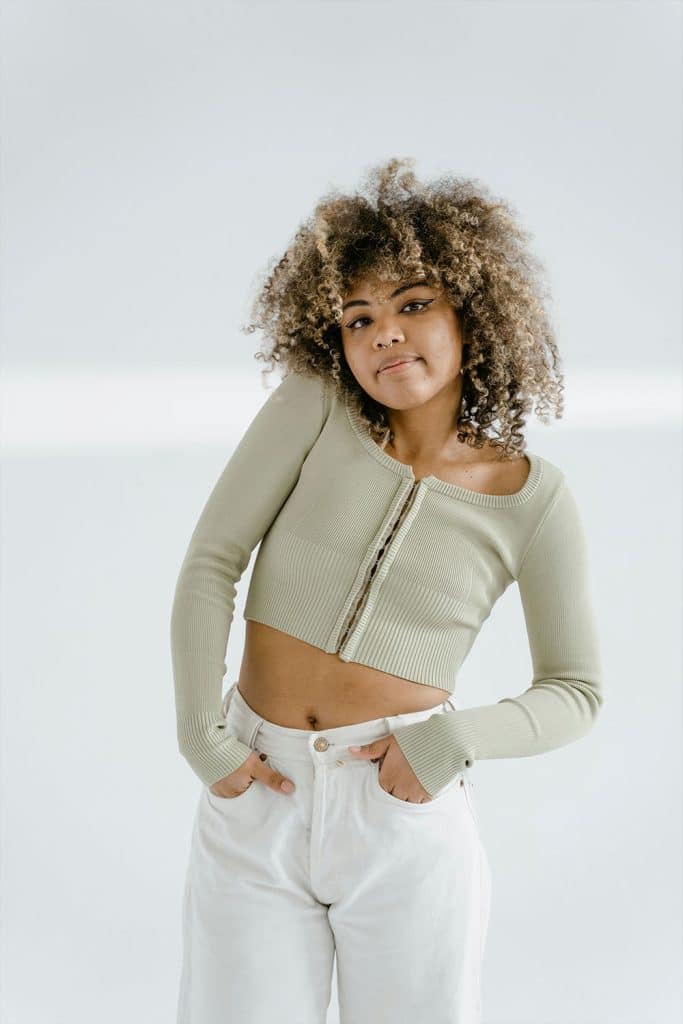 Woman with curly hair wearing a cropped sweater and white pants, hands in pockets.