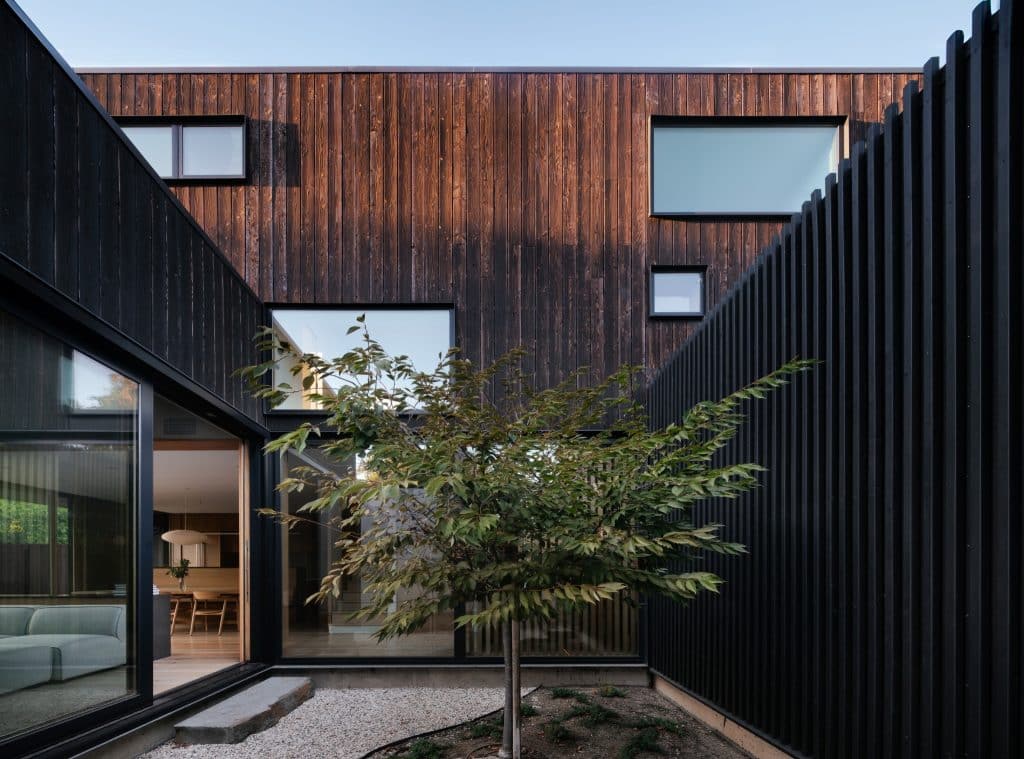 Modern house with dark wood exterior, large windows, and a small tree in a gravel courtyard.