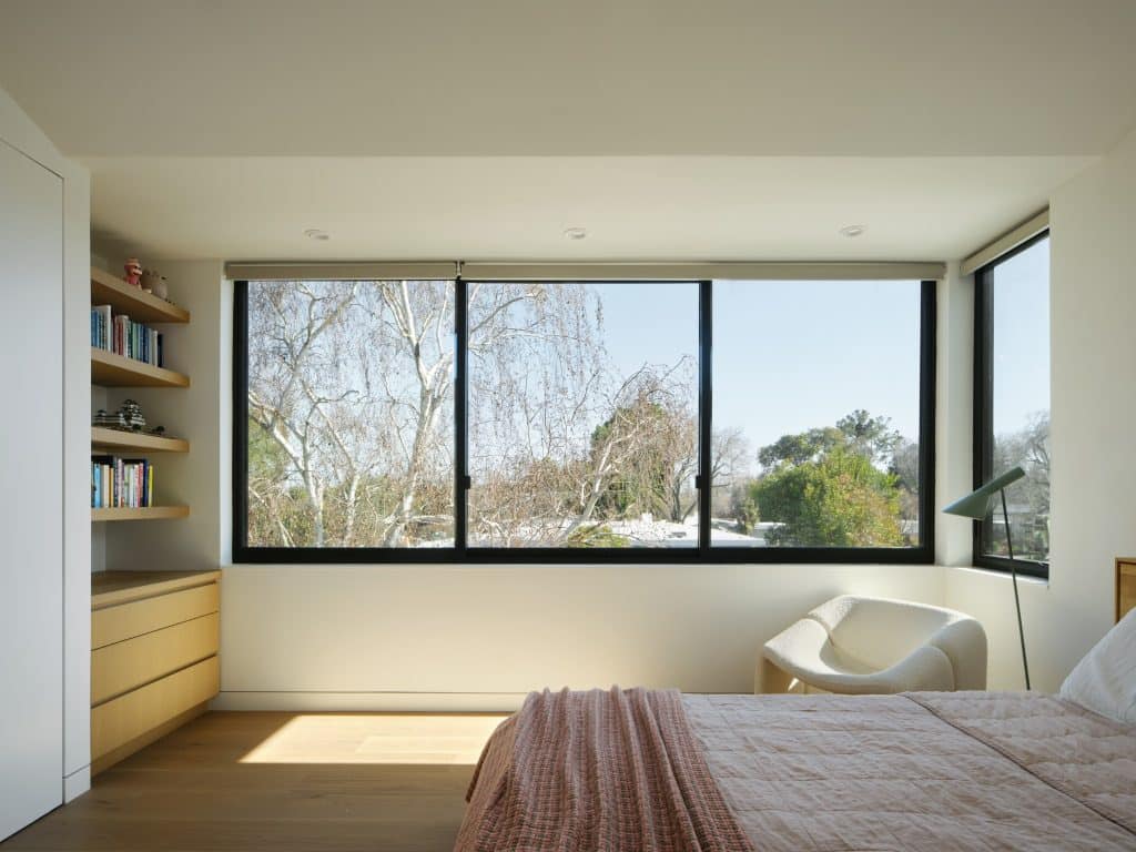 Minimalist bedroom with large windows, a cozy chair, and shelves with books and drawers.