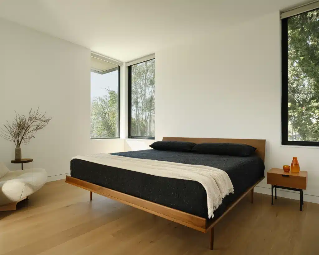 Minimalist bedroom with a wooden bed, black bedding, large windows, and light wood floors.