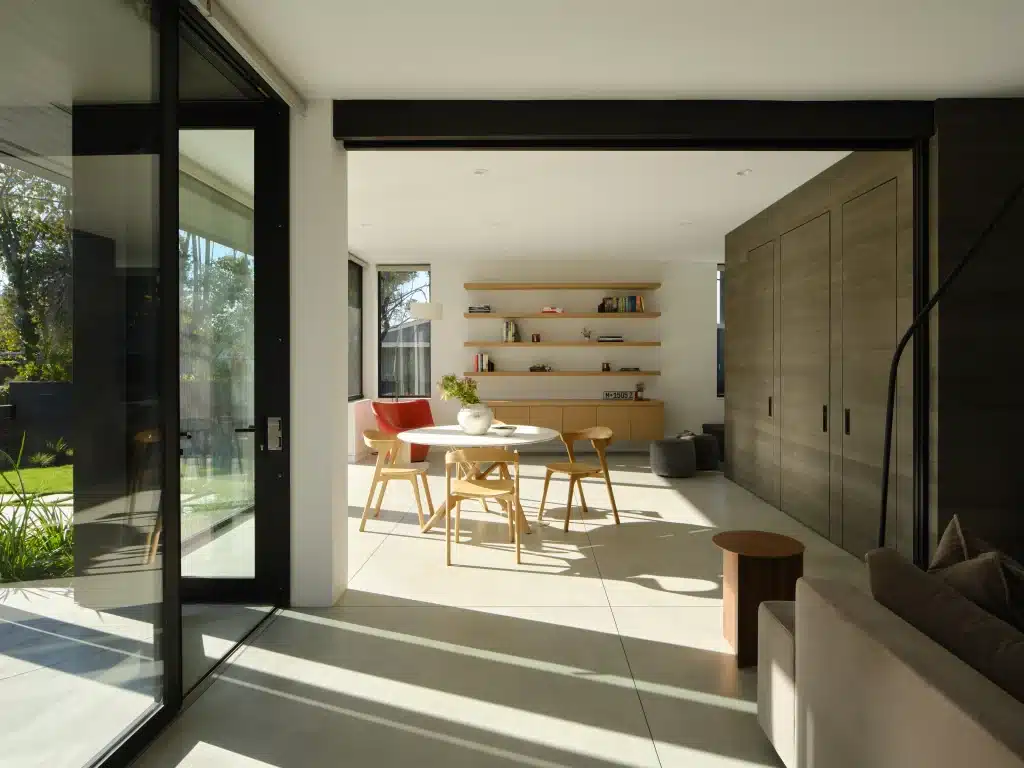 Modern dining area with wooden table and chairs, sunlit through large glass doors.