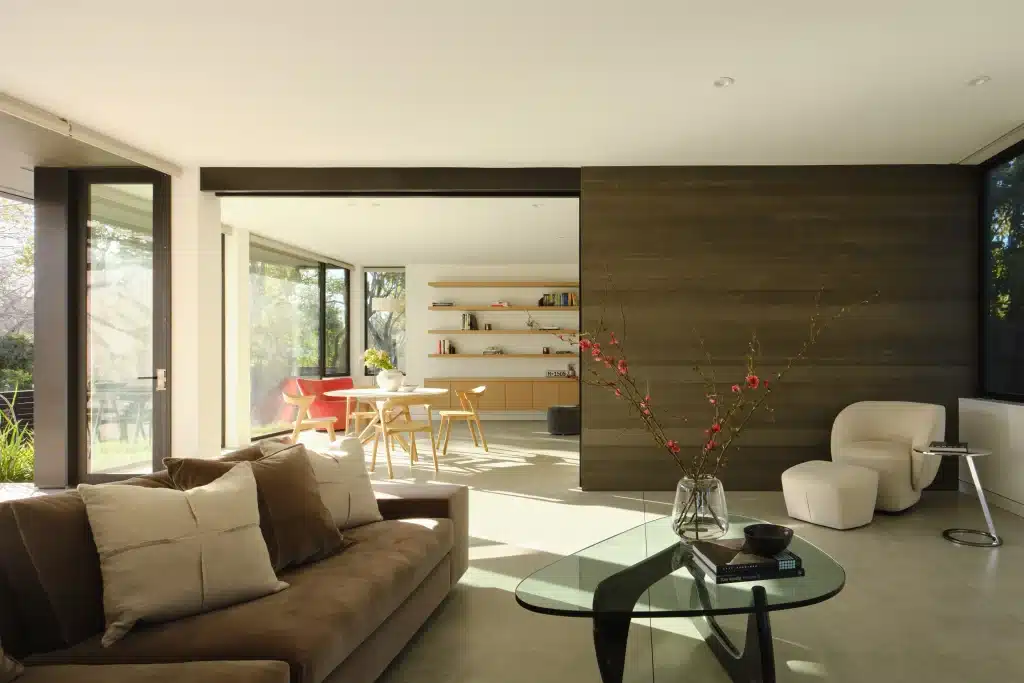 Modern living room with brown sofa, glass coffee table, and bright dining area in background.