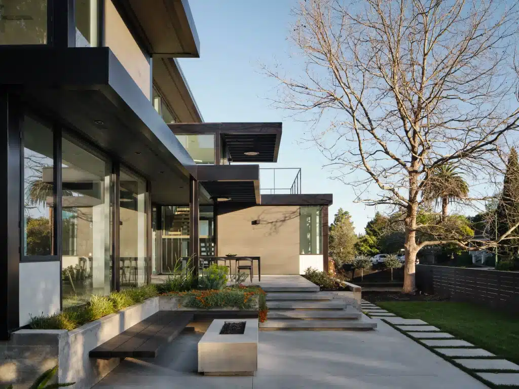 Modern house with large glass windows, patio seating, and a tree with no leaves nearby.