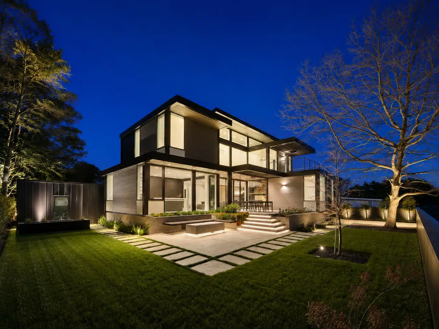 Modern two-story house with large windows, lit up at night, and a landscaped yard.