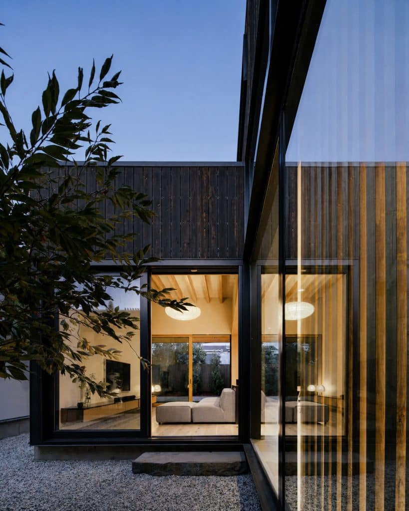 Modern house exterior with large glass windows glowing warmly at dusk, tree in foreground.