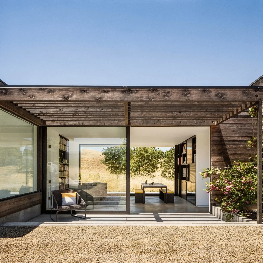 Modern glass room with a wood pergola, desk, chair, and outdoor plants under clear sky.