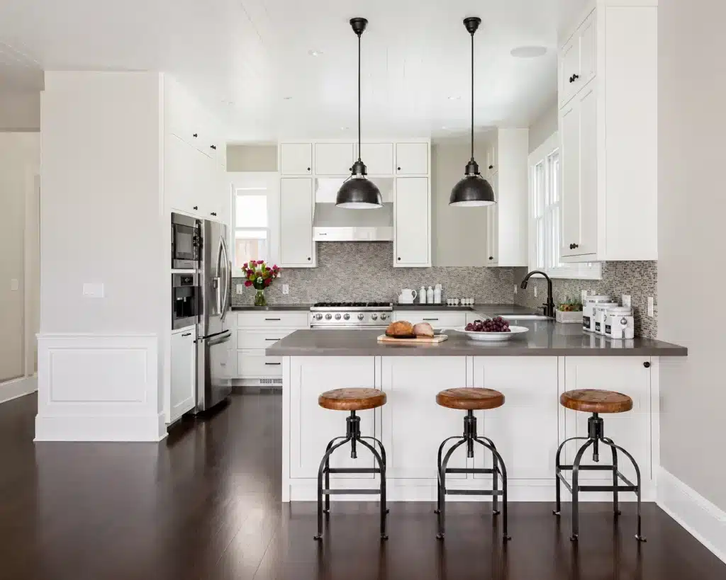 Modern white kitchen with island, pendant lights, and three wooden stools on dark flooring.
